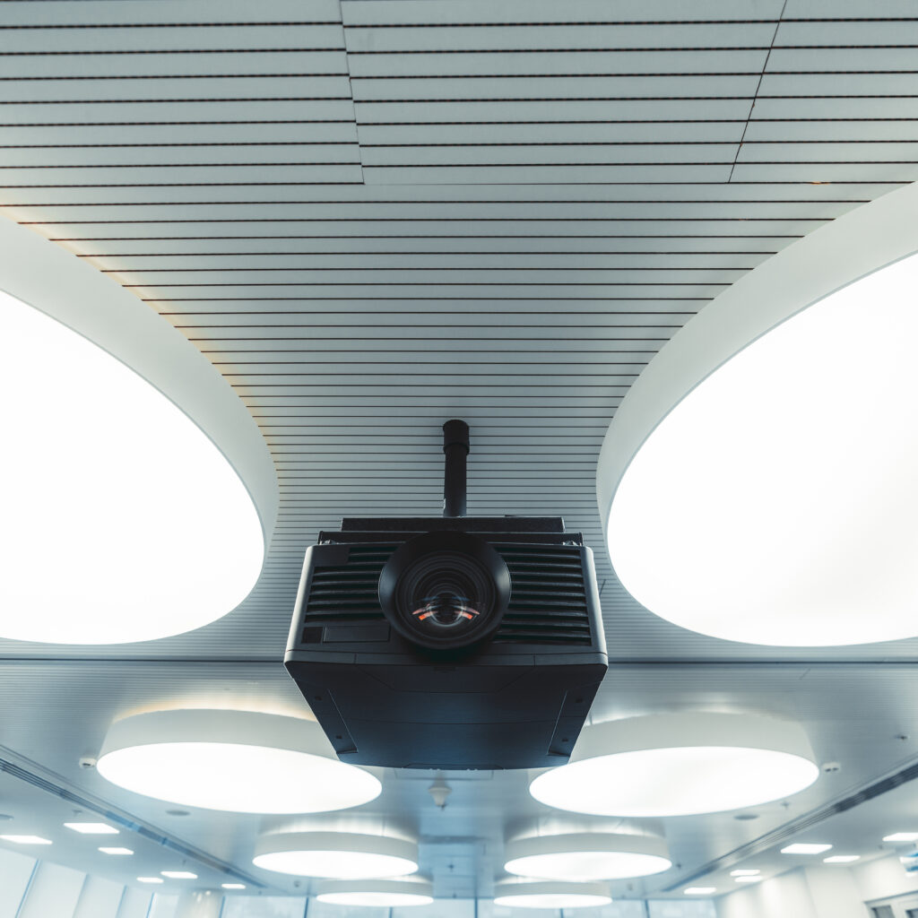 Wide-angle view of a black modern projector with a plastic case and a huge lens with aberrations on it, mounted to the ceiling with multiple huge round ceiling lamps lighting a modern conference room