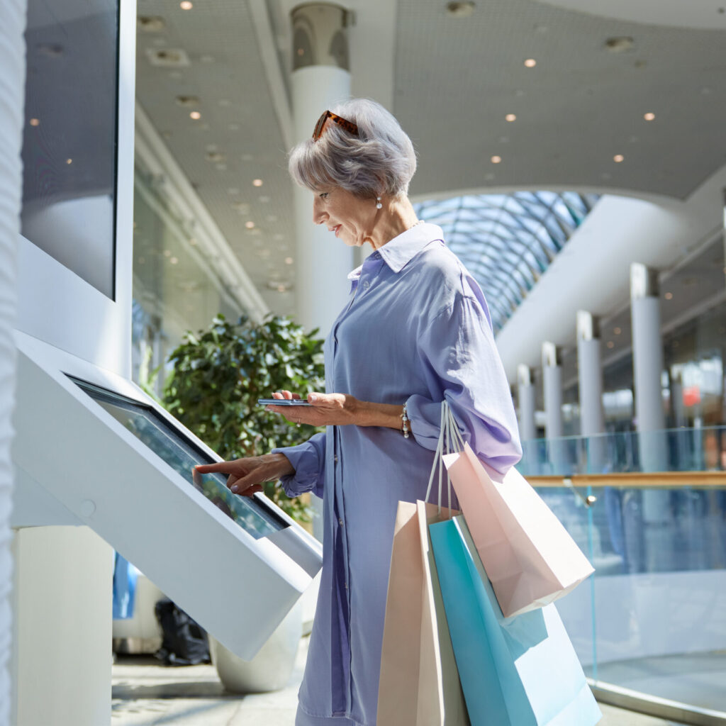 Senior old woman using atm machine for money cash withdrawing in shopping mall. Stylish elderly lady holding bags with purchases standing front of digital self-service kiosk